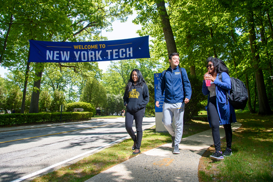 Three students walk near a Welcome to New York Tech sign outdoors on the Long Island Campus.